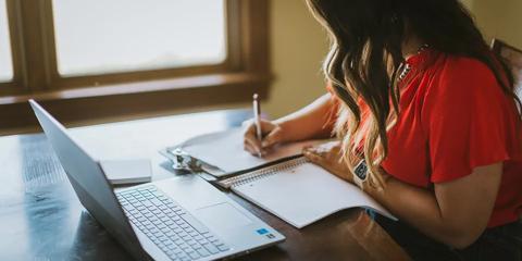 woman sitting at a desk writing in a notebook and looking at a laptop