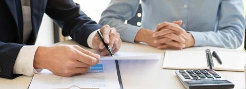 Two people sitting at a table, one holding a pen and pointing at a piece of paper, the other with his hands folded. A notebook and calculator also sit on the table