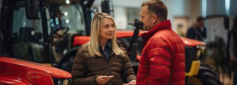 A man and a woman talking in a shed with red tractors behind them