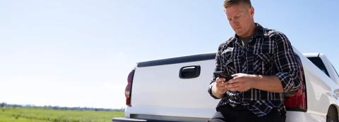 farmer looking at phone while leaning against tailgate of white pickup truck in a rural setting
