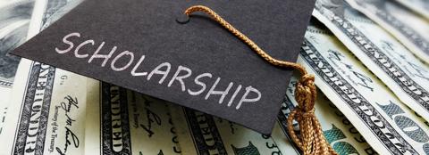 Close up of a graduation cap with the word 'Scholarship' written on it and $100 bills in the background