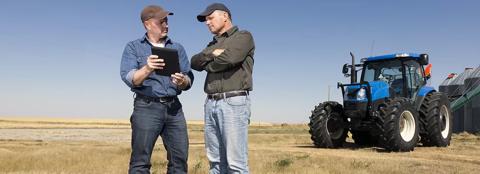 Two men in a field looking at a tablet with a tractor in the background