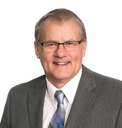 A portrait of a man wearing a gray suit, white shirt, and blue patterned tie with glasses