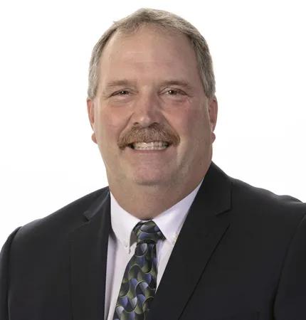 A portrait of a man with a mustache wearing a black jacket, white shirt, and green/blue patterned tie